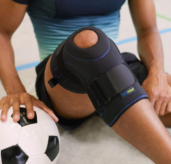 Muškarac koji nosi steznik za koljeno sjedi na podu i stavlja ruku na nogometnu loptu Muškarac koji nosi steznik za koljeno sjedi na podu i stavlja ruku na nogometnu loptu